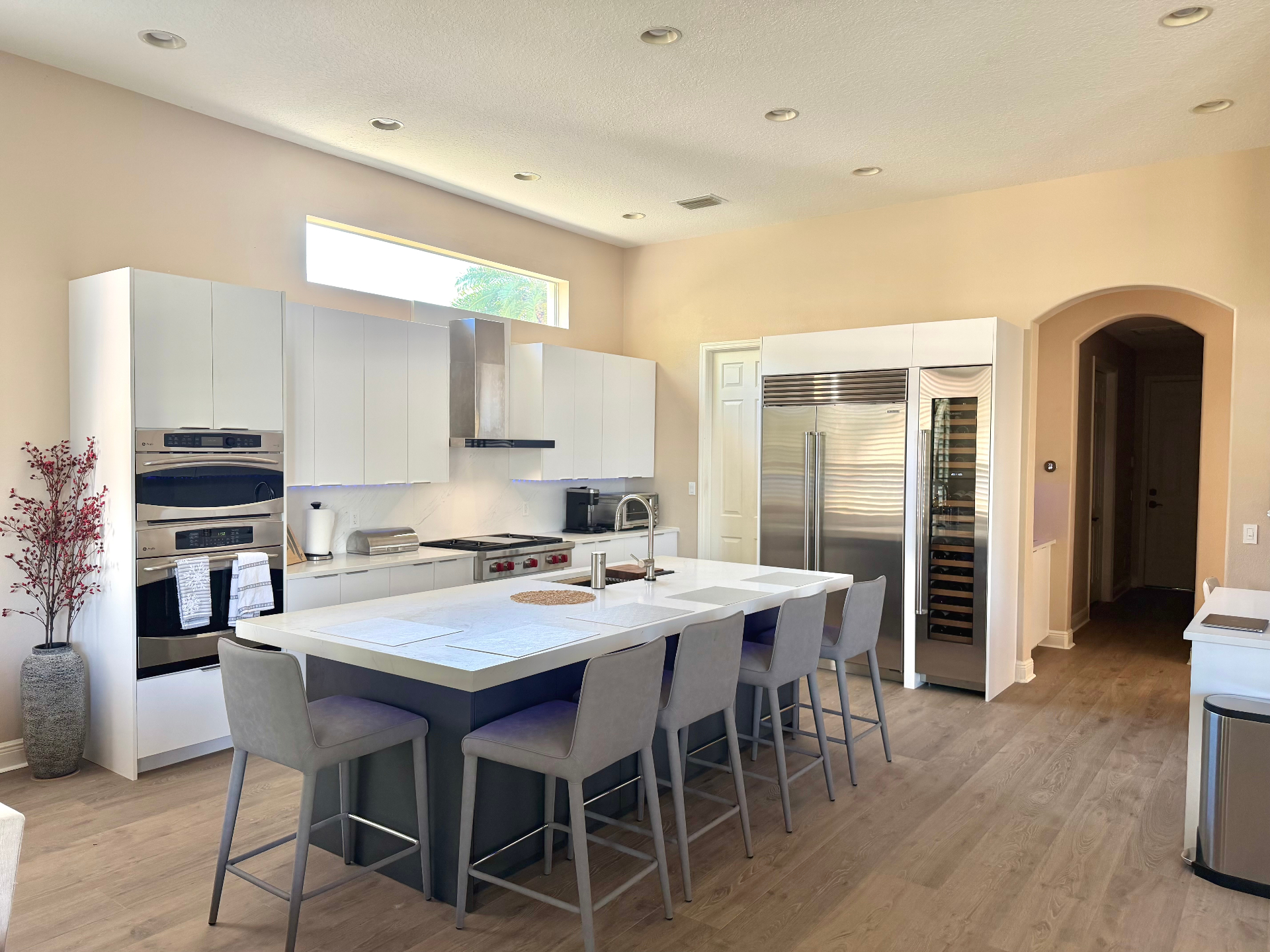 Modern white kitchen remodel with large waterfall island, bar seating, double ovens, and built-in wine fridge